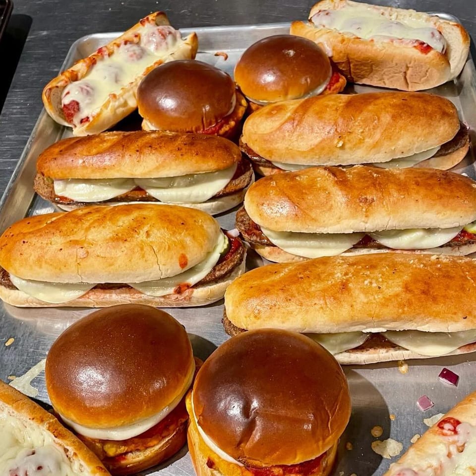A metal tray filled with various Lucy Blue Pizza sandwiches, including meatball subs and cheeseburger sliders.