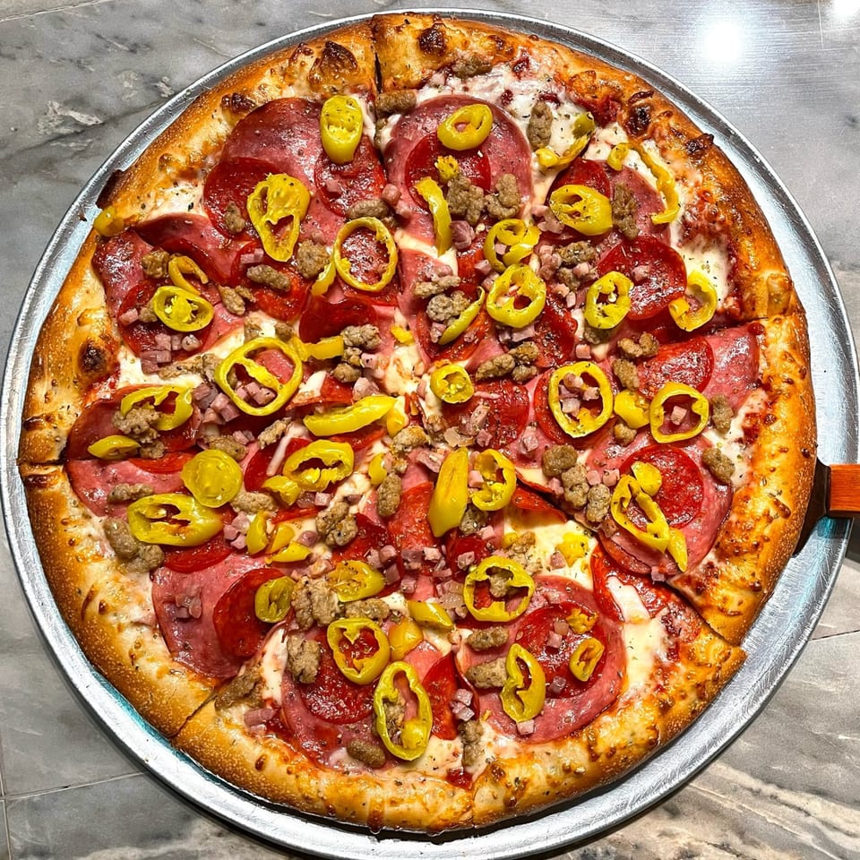 A Lucy Blue Pizza with pepperoni, sausage, ham, and yellow banana peppers on a metal tray.